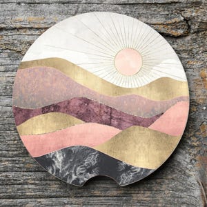 May include: Round car coaster with a landscape design. The coaster features a sun with radiating lines, and layered hills in shades of pink, gold, and purple. The bottom section is a dark gray and white pattern.