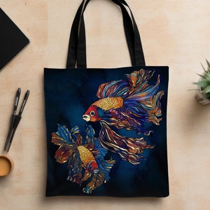 May include: A tote bag with a dark blue background featuring two colorful betta fish illustrations. The fish have intricate patterns in shades of blue, orange, and gold. The bag has black handles and is placed on a light brown surface.