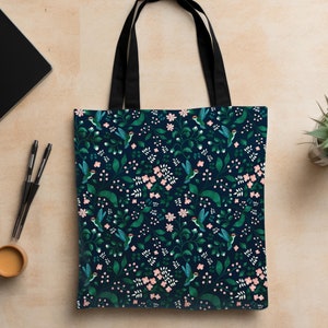 May include: A black canvas tote bag with black straps. The bag features a repeating pattern of green leaves, pink flowers, and blue hummingbirds on a dark blue background.