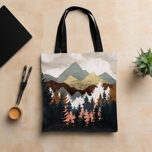 May include: A tote bag with a mountain landscape design. The bag features a forest of trees in shades of black, brown, and peach, with mountains in the background. The bag has black handles and is made of a sturdy material.