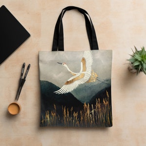 May include: A tote bag with a white crane flying over a dark blue and green landscape with gold accents. The crane is flying towards the right side of the bag. The bag has black straps.