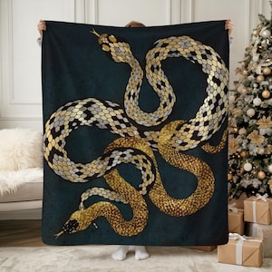 May include: A dark teal blanket featuring a detailed illustration of two intertwined snakes. The snakes have gold, black, and silver scales, with their bodies forming elegant curves against the dark background. The blanket is held up by a person.