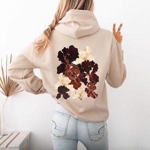 May include: Beige hoodie featuring a floral design on the back. The graphic showcases a cluster of irises in shades of burgundy, brown, and cream. The hoodie has a drawstring hood and long sleeves, offering a casual and comfortable style.