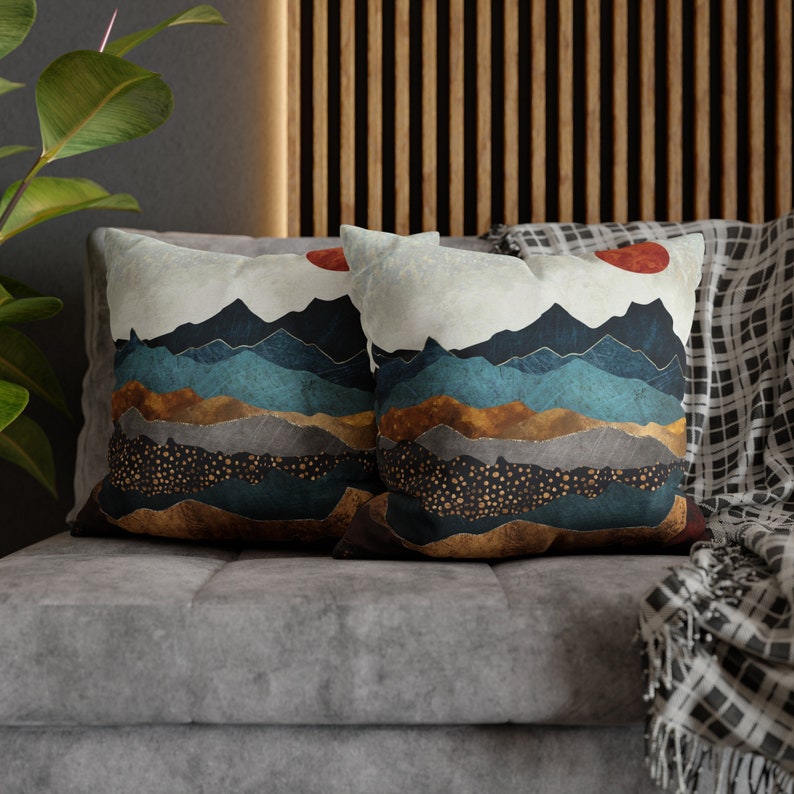May include: Two decorative throw pillows with a mountain landscape design. The pillows feature a white background with blue, brown, and gold mountains, a red sun, and gold glitter accents.