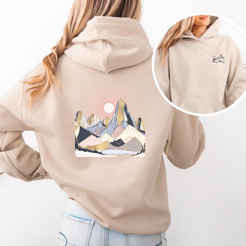Pu&ograve; includere: Felpa con cappuccio beige con grafica di una catena montuosa e un sole nascente. Il design include sfumature di blu, rosa, oro e grigio. La felpa ha una tasca frontale e un cappuccio con coulisse. L'immagine mostra il retro e una vista pi&ugrave; piccola del davanti.