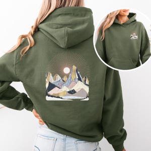 Pu&ograve; includere: Felpa con cappuccio verde oliva con grafica di catena montuosa sul retro e una grafica di montagna pi&ugrave; piccola sul davanti. La grafica della montagna presenta un sole, cime e vari colori tra cui blu, rosa e oro.