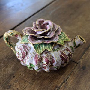 May include: A decorative teapot with a rose-shaped lid. The teapot is primarily light pink with burgundy and gold accents. The rose lid is a darker shade of pink with green leaves and gold trim. The handle and spout are also detailed with gold.
