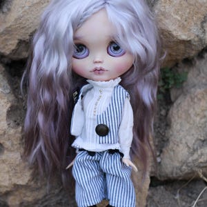May include: A doll with long, wavy, light purple hair and large, light purple eyes. The doll wears a white high-neck blouse with small buttons, a blue and white pinstriped vest with a dark brown button, matching pinstriped shorts, and black shoes.