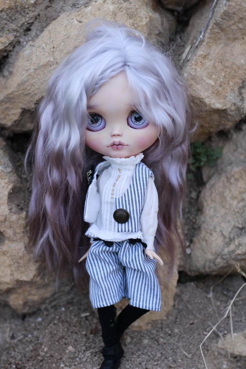 May include: A doll with long, wavy, light purple hair and large, light purple eyes. The doll wears a white high-neck blouse with small buttons, a blue and white pinstriped vest with a dark brown button, matching pinstriped shorts, and black shoes.