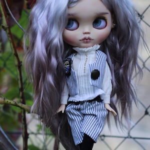 May include: A doll with long, wavy, light purple hair and large blue eyes. The doll wears a white high-neck shirt, a blue and white pinstriped vest with a dark button, matching pinstriped shorts, black tights, and black shoes with straps.