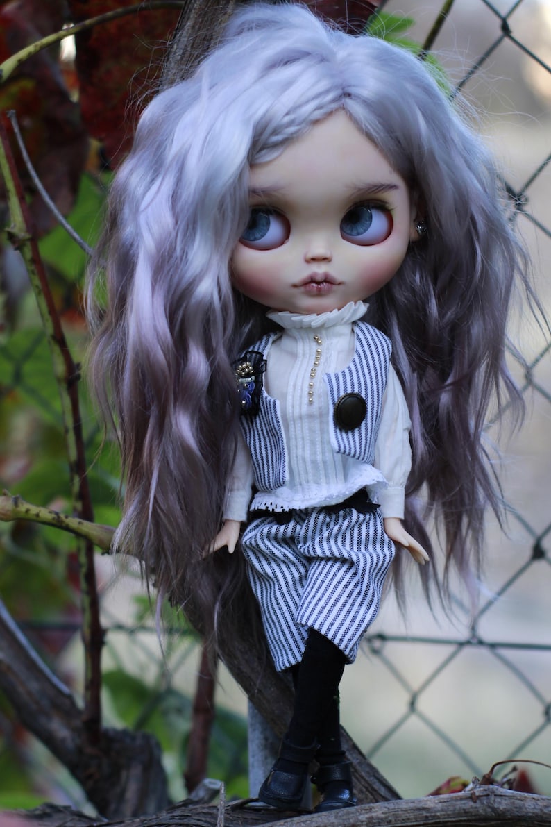 May include: A doll with long, wavy, light purple hair and large blue eyes. The doll wears a white high-neck shirt, a blue and white pinstriped vest with a dark button, matching pinstriped shorts, black tights, and black shoes with straps.