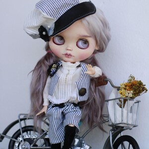 May include: A doll with long lavender hair wearing a white and blue striped newsboy cap, a white ruffled blouse, a matching striped vest, and striped shorts. The doll is positioned next to a small metal bicycle with a basket containing dried flowers.