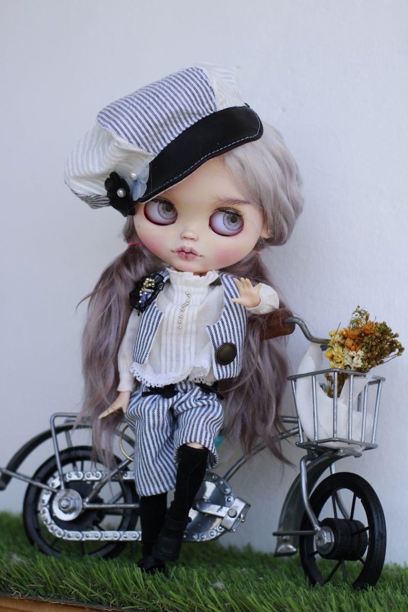 May include: A doll with long lavender hair wearing a white and blue striped newsboy cap, a white ruffled blouse, a matching striped vest, and striped shorts. The doll is positioned next to a small metal bicycle with a basket containing dried flowers.