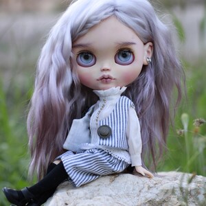 May include: A doll with long, wavy, light purple hair and large blue eyes. The doll wears a white collared shirt with a blue and white striped vest and matching shorts, black tights, and black shoes. The doll has small silver stud earrings.