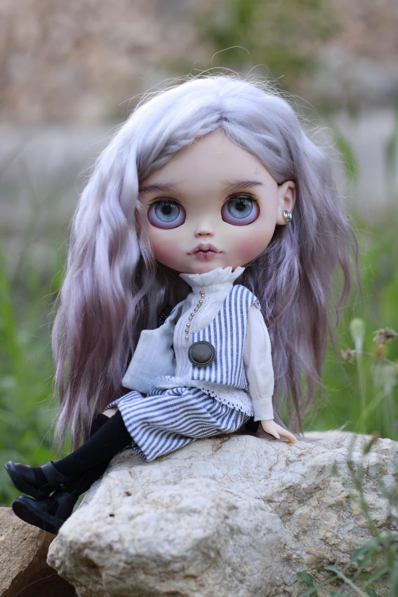 May include: A doll with long, wavy, light purple hair and large blue eyes. The doll wears a white collared shirt with a blue and white striped vest and matching shorts, black tights, and black shoes. The doll has small silver stud earrings.