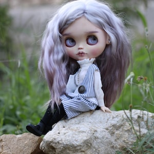 May include: A doll with long wavy lavender hair, large grey eyes, and pale skin. The doll wears a white high-neck blouse under a blue and white pinstriped vest and matching skirt, paired with black tights and black shoes with small buckles.