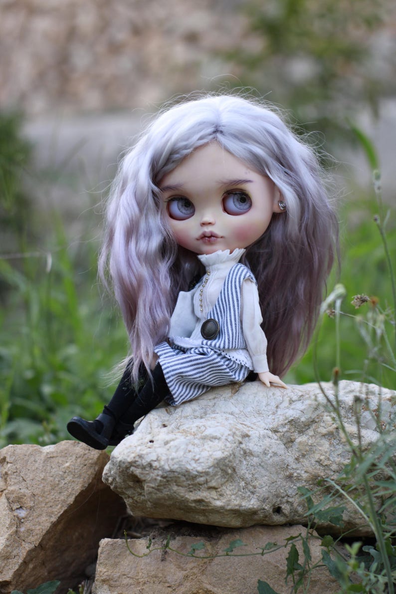 May include: A doll with long wavy lavender hair, large grey eyes, and pale skin. The doll wears a white high-neck blouse under a blue and white pinstriped vest and matching skirt, paired with black tights and black shoes with small buckles.