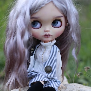 May include: A doll with long wavy lavender hair and large blue eyes. The doll wears a white high-neck blouse with small gold buttons, a blue and white pinstriped vest, black leggings, and black shoes. The doll has a pale complexion and pink lips.