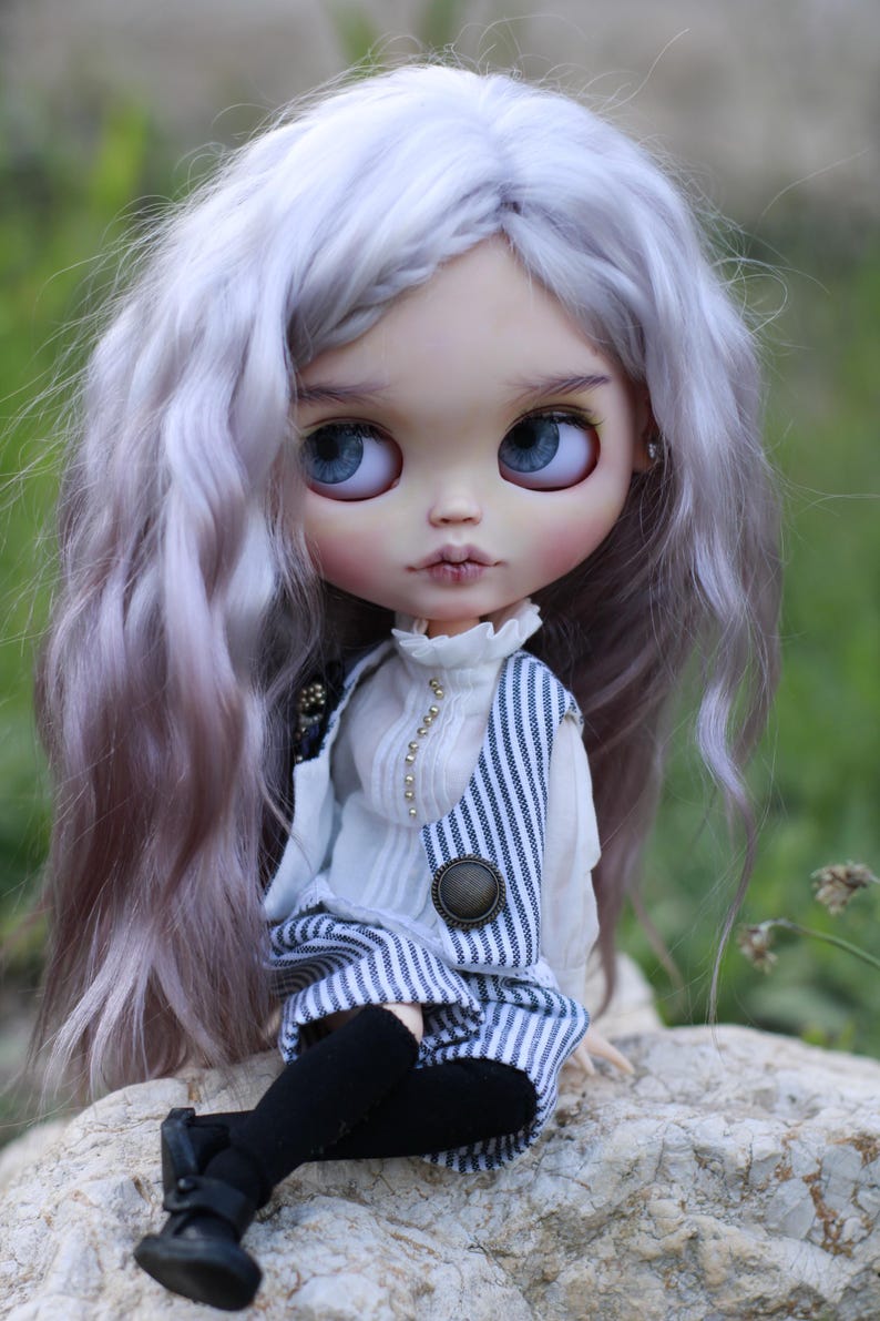 May include: A doll with long wavy lavender hair and large blue eyes. The doll wears a white high-neck blouse with small gold buttons, a blue and white pinstriped vest, black leggings, and black shoes. The doll has a pale complexion and pink lips.