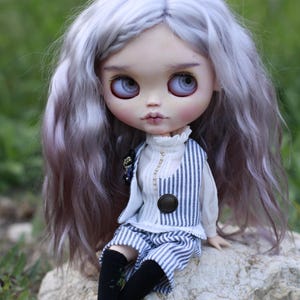 May include: A doll with long wavy lavender hair, large light blue eyes, and pale skin. The doll wears a white high-neck blouse under a blue and white pinstriped vest and matching shorts, paired with black knee-high socks and black shoes.