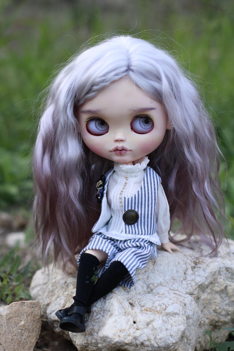 May include: A doll with long wavy lavender hair, large light blue eyes, and pale skin. The doll wears a white high-neck blouse under a blue and white pinstriped vest and matching shorts, paired with black knee-high socks and black shoes.