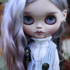 May include: A doll with long, wavy, light lavender hair and large, pale blue eyes. The doll wears a white high-neck blouse with a vertical row of small beads, paired with a blue and white pinstriped vest featuring a single round bronze button.
