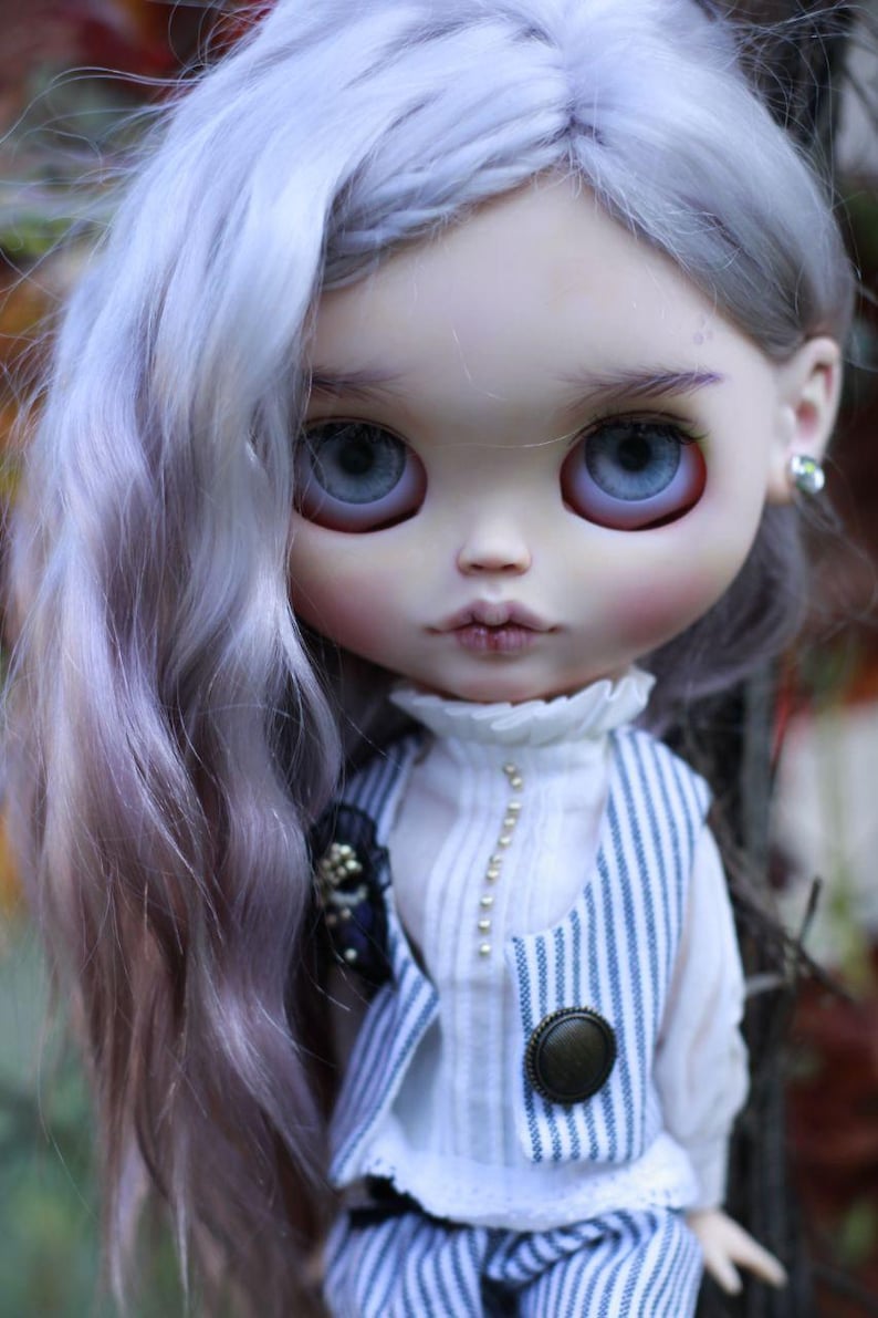 May include: A doll with long, wavy, light lavender hair and large, pale blue eyes. The doll wears a white high-neck blouse with a vertical row of small beads, paired with a blue and white pinstriped vest featuring a single round bronze button.