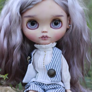 May include: A doll with long, wavy, light lavender hair and large, detailed eyes. The doll wears a white high-neck blouse with small gold buttons, a blue and white pinstriped vest with a large round button, matching pinstriped shorts, and black socks.