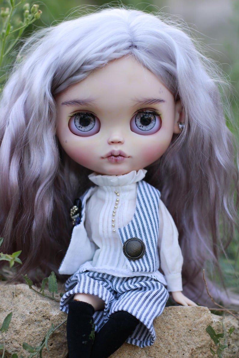 May include: A doll with long, wavy, light lavender hair and large, detailed eyes. The doll wears a white high-neck blouse with small gold buttons, a blue and white pinstriped vest with a large round button, matching pinstriped shorts, and black socks.