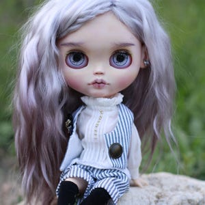 May include: A doll with long wavy lavender hair, large grey eyes, and pale skin. The doll wears a white high-neck blouse with a vertical row of small beads, a blue and white pinstriped vest with a round button, matching pinstriped shorts, and black socks.