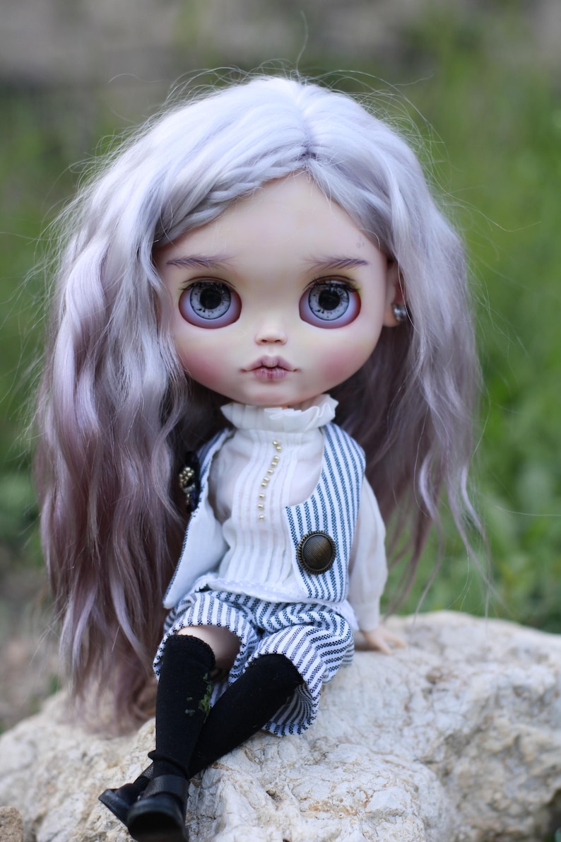 May include: A doll with long wavy lavender hair, large grey eyes, and pale skin. The doll wears a white high-neck blouse with a vertical row of small beads, a blue and white pinstriped vest with a round button, matching pinstriped shorts, and black socks.