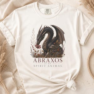 以下が含まれることがあります： クリーム色の半袖Tシャツで、野生の花々に囲まれたダークなドラゴンのグラフィックが描かれています。イラストの下には、ダークなセリフ体で「ABRAXOS IS MY SPIRIT ANIMAL」というテキストがプリントされています。Tシャツにはクルーネックとラベルが付いています。