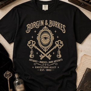 May include: A black short-sleeve t-shirt featuring a gold graphic print. The design includes an eye inside an ornate oval frame, two crossed keys with small bottles hanging from them, and the text Borgin & Burkes, Antiques, Oddities, Dark Artefacts, Knockturn Alley, and Est. 1863.
