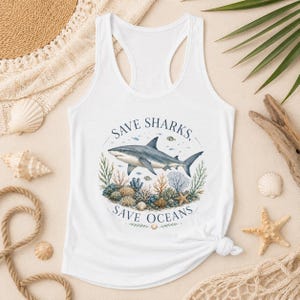 Op de afbeelding: Een wit racerback-hemd met een cirkelvormige afbeelding van een haai die boven een koraalrif zwemt. De tekst "SAVE SHARKS, SAVE OCEANS" is in een donkerblauw lettertype in een boog rond de centrale illustratie van de haai en het rif gedrukt.