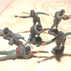 May include: A collection of vintage toy soldiers in various poses, including kneeling, standing, and prone positions. The soldiers are painted in olive green uniforms with black helmets and are holding rifles and grenades. The toys are on a light brown surface.