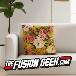 Puede incluir: Cojín cuadrado amarillo mostaza con un estampado floral de rosas rosas, rojas y blancas, y hojas verdes. El cojín está sobre un sofá blanco. El texto "THE FUSION GEEK.COM" está en la parte inferior.