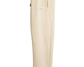 May include: Beige sweatpants with a white stripe at the waistband and a zippered pocket on the right leg. The sweatpants have the word "SERENITI" embroidered on the left leg.