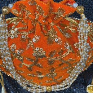 May include: An orange velvet pouch with gold embroidery and sequins. The drawstring bag features a beaded tassel and a gold beaded handle. The bag is a vibrant accessory.
