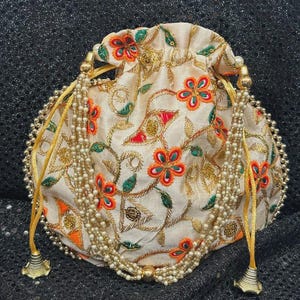 May include: Cream drawstring pouch with floral embroidery in orange, red, and green. The bag has gold accents, including a beaded trim and tassel details. A stylish accessory for special occasions.