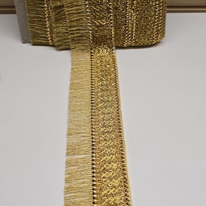 May include: A spool of metallic gold fringe trim featuring a woven lattice pattern along the centre and straight, fine gold threads extending from one side. The trim is wound around a rectangular cardboard core, showing a continuous length.
