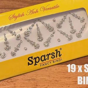 May include: A yellow box with a clear window showing 19 silver bindis. The bindis are in various shapes and sizes, with a sparkling, rhinestone-like appearance. The box has the text "Stylish Anh Versatile" and "Sparsh Fancy Bindi".