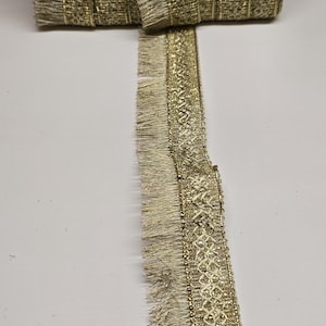 May include: A roll of metallic gold trim featuring a woven lattice pattern and a long, straight fringe edge. The decorative ribbon has a shimmering finish and is displayed as a continuous strip extending from the main spool of fabric trim.