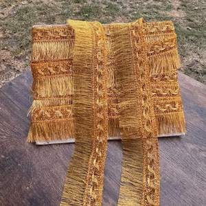 May include: A roll of metallic gold fringe trim featuring a decorative woven border with a repeating wave pattern. The trim consists of a wide band of dense, straight golden threads hanging vertically from a structured, textured fabric header.
