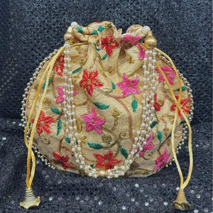 May include: A gold drawstring pouch featuring red and pink floral embroidery. The pouch has a beaded trim and a pearl-like beaded strand. The drawstring cords end in gold bell-shaped tassels. The pouch is approximately 15 cm in diameter.