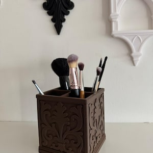 Op de afbeelding: Een bruine, vierkante make-up kwastenhouder met een sierlijk, verhoogd bloemmotief. De houder is verdeeld in compartimenten en bevat diverse make-up kwasten met zwarte, roze en bruine handvatten en borstelharen. Achtergrond is een witte muur met decoratieve accenten.