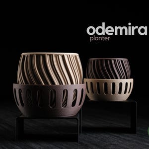 Può includere: Due fioriere decorative dal design unico. La fioriera più grande è beige e marrone, mentre quella più piccola è marrone e beige. Entrambe le fioriere hanno un motivo a spirale e poggiano su una base marrone scuro. Vengono visualizzate le parole "odemira" e "planter".