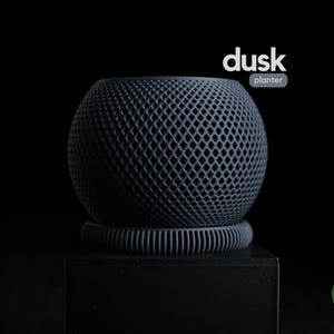 Könnte beinhalten: Ein dunkelgrauer, kugelförmiger Pflanztopf mit Gittermuster, der auf einem passenden Sockel steht. Das Wort "dusk" steht über dem Wort "planter" in weißem Text. Der Pflanztopf steht auf einer schwarzen Oberfläche.