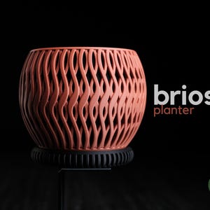 May include: A terracotta-colored planter with a woven design, sitting on a black base. The planter has the word "brios" and "planter" in white and orange text. The background is black.