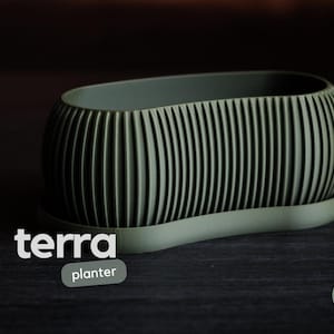 Könnte beinhalten: Ein olivgrüner Pflanztopf mit geripptem Design, der auf einer passenden Untertasse steht. Das Wort "terra" wird in weißem Text angezeigt, darunter "planter". Das Design stammt von h3li0 design.
