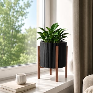 Puede incluir: Una maceta cilíndrica negra con textura de estrías verticales, sostenida por un soporte de madera de cuatro patas. La maceta contiene una planta verde frondosa con follaje puntiagudo. El conjunto se sitúa sobre una superficie blanca.